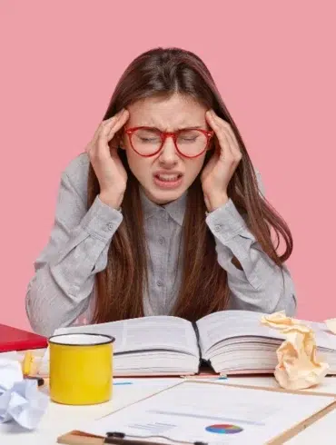 Student with red-rimmed glasses holding head in hands with open book and crumpled papers on desk.
