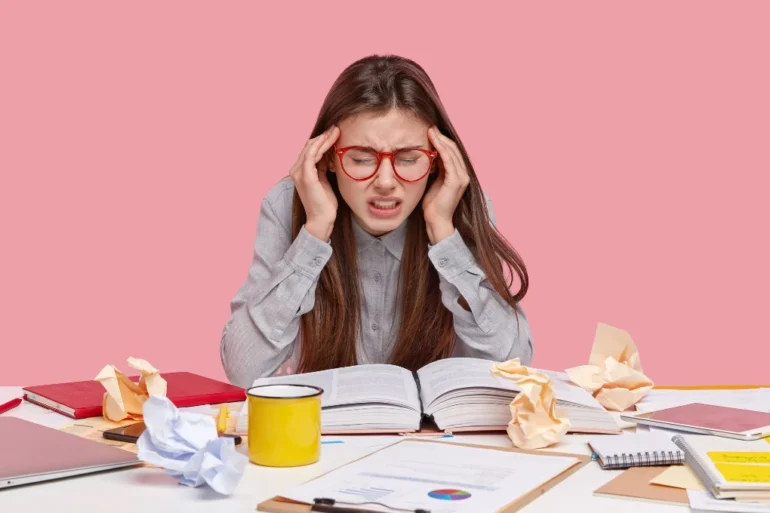 Student with red-rimmed glasses holding head in hands with open book and crumpled papers on desk.