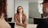 A woman in glasses and a beige blazer sits confidently at a table, smiling at two colleagues during a meeting in a modern office. The atmosphere is professional.