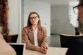 A woman in glasses and a beige blazer sits confidently at a table, smiling at two colleagues during a meeting in a modern office. The atmosphere is professional.