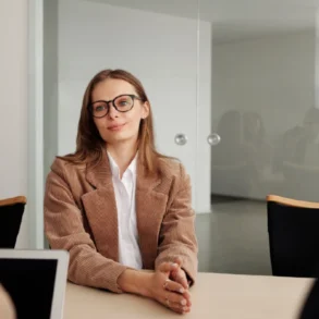 A woman in glasses and a beige blazer sits confidently at a table, smiling at two colleagues during a meeting in a modern office. The atmosphere is professional.