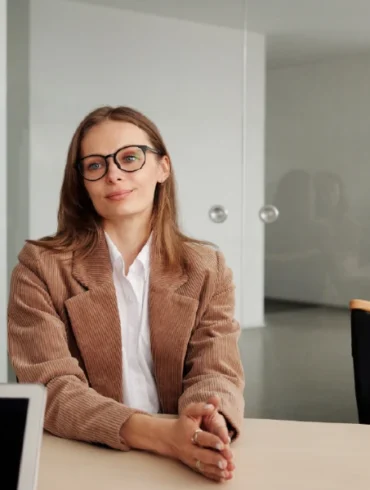 A woman in glasses and a beige blazer sits confidently at a table, smiling at two colleagues during a meeting in a modern office. The atmosphere is professional.
