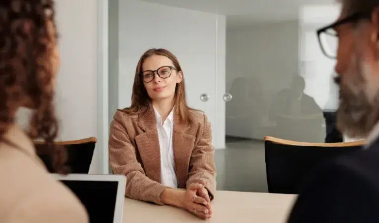 A woman in glasses and a beige blazer sits confidently at a table, smiling at two colleagues during a meeting in a modern office. The atmosphere is professional.