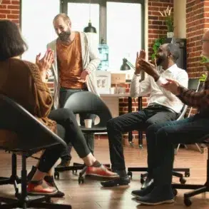 A diverse group of four people sits in a cozy room with brick walls, clapping and smiling warmly at a man standing in the center. The mood is positive and supportive.