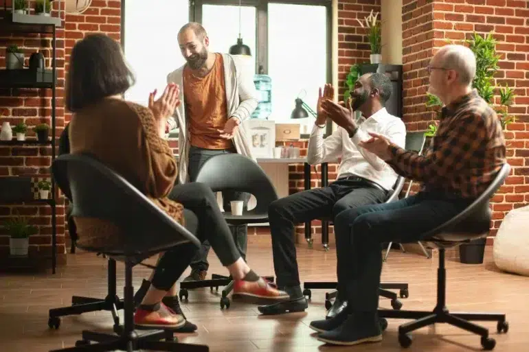 A diverse group of four people sits in a cozy room with brick walls, clapping and smiling warmly at a man standing in the center. The mood is positive and supportive.