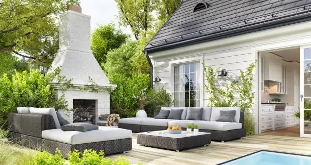 Modern patio with wicker sofas, cushions, and a coffee table beside a pool. A white outdoor fireplace is surrounded by lush greenery, creating a serene atmosphere.