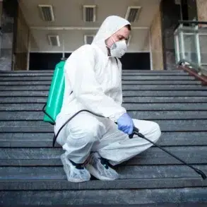 A person in protective gear, including a mask, kneels on stairs indoors, spraying disinfectant from a green backpack sprayer. The setting is calm and serious.