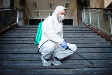 A person in protective gear, including a mask, kneels on stairs indoors, spraying disinfectant from a green backpack sprayer. The setting is calm and serious.