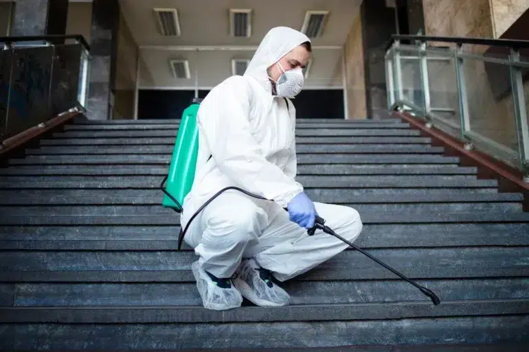 A person in protective gear, including a mask, kneels on stairs indoors, spraying disinfectant from a green backpack sprayer. The setting is calm and serious.