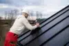 A worker in a white hard hat and red pants installs solar panels on a metal roof. Overcast sky and trees in the background; focused and industrious mood.