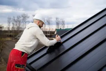 A worker in a white hard hat and red pants installs solar panels on a metal roof. Overcast sky and trees in the background; focused and industrious mood.
