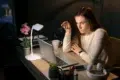 A woman in a white sweater sits at a desk, looking intently at a laptop. She holds eyeglasses in one hand. The scene is warmly lit by a desk lamp. Nearby are a potted orchid, coffee cup, and headphones, suggesting focus and concentration.