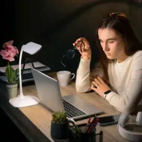 A woman in a white sweater sits at a desk, looking intently at a laptop. She holds eyeglasses in one hand. The scene is warmly lit by a desk lamp. Nearby are a potted orchid, coffee cup, and headphones, suggesting focus and concentration.