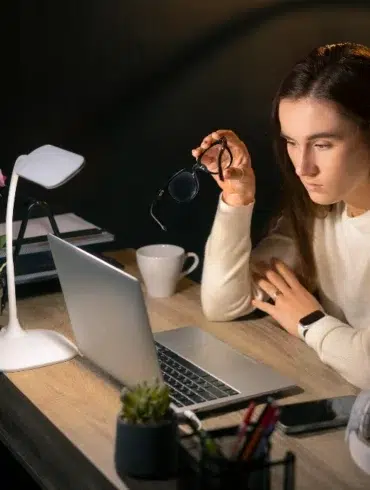 A woman in a white sweater sits at a desk, looking intently at a laptop. She holds eyeglasses in one hand. The scene is warmly lit by a desk lamp. Nearby are a potted orchid, coffee cup, and headphones, suggesting focus and concentration.
