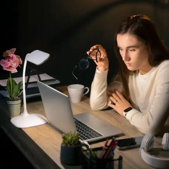 A woman in a white sweater sits at a desk, looking intently at a laptop. She holds eyeglasses in one hand. The scene is warmly lit by a desk lamp. Nearby are a potted orchid, coffee cup, and headphones, suggesting focus and concentration.