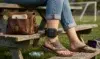 A person wearing jeans and sandals sits on a wooden bench in a park, with an ankle monitor visible. A leather bag rests nearby. The mood is casual.