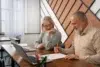 Older man and woman sit at a wooden table with laptops, smiling and collaborating over a drawing. The room has geometric wood paneling.