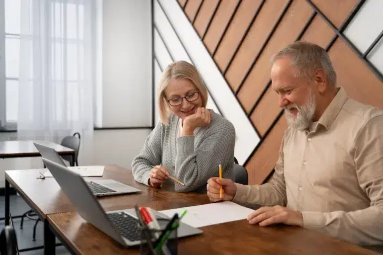 Older man and woman sit at a wooden table with laptops, smiling and collaborating over a drawing. The room has geometric wood paneling.