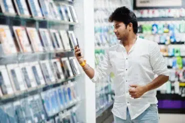 Man in a white shirt looking at a smartphone in a store aisle filled with various electronic gadgets and accessories.