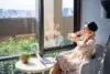 Woman wearing a black fedora and a polka dot dress sits by a window with a laptop and a plant.