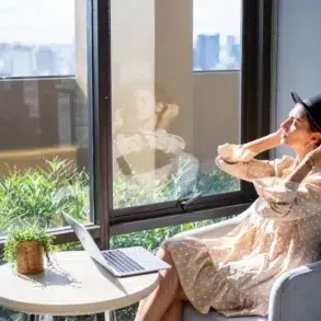 Woman wearing a black fedora and a polka dot dress sits by a window with a laptop and a plant.