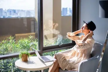 Woman wearing a black fedora and a polka dot dress sits by a window with a laptop and a plant.