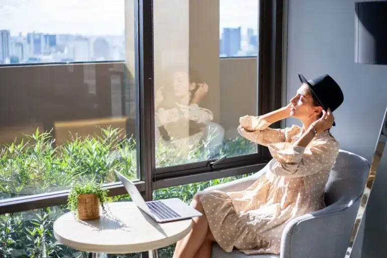 Woman wearing a black fedora and a polka dot dress sits by a window with a laptop and a plant.