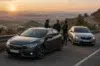 Three men stand beside a dark gray sedan and a silver sedan parked on a scenic mountain road at sunset.