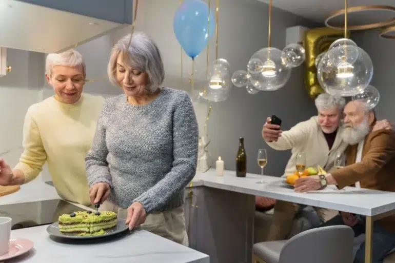 Two senior women cut a cake in a kitchen decorated with balloons and modern lights, while two senior men take a selfie in the background.