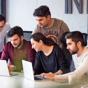 A group of five colleagues collaborate around laptops in a modern office. They appear focused and engaged, sharing ideas in a team discussion.