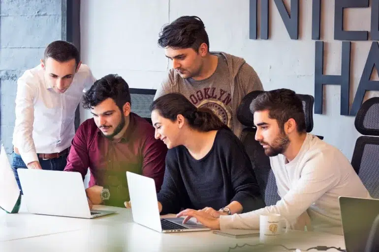 A group of five colleagues collaborate around laptops in a modern office. They appear focused and engaged, sharing ideas in a team discussion.