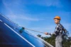 A person in a hard hat cleaning solar panels with a brush under a clear blue sky, emphasizing renewable energy and maintenance work.