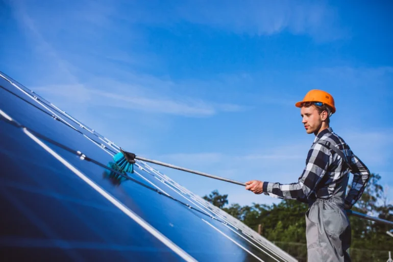 A person in a hard hat cleaning solar panels with a brush under a clear blue sky, emphasizing renewable energy and maintenance work.