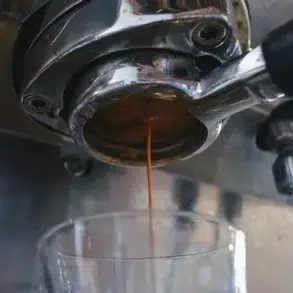 Fresh espresso pours from a coffee machine's portafilter into a clear glass below.