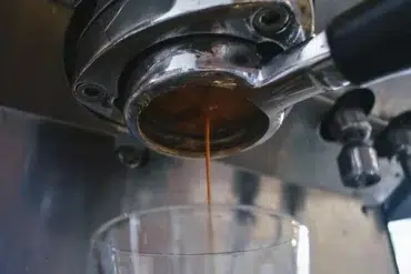 Fresh espresso pours from a coffee machine's portafilter into a clear glass below.