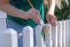 Close-up of a person painting a white picket fence with a brush, adding green paint. They wear a teal shirt, conveying a calm, creative mood.
