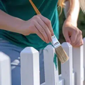 Close-up of a person painting a white picket fence with a brush, adding green paint. They wear a teal shirt, conveying a calm, creative mood.