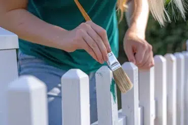 Close-up of a person painting a white picket fence with a brush, adding green paint. They wear a teal shirt, conveying a calm, creative mood.