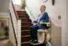 Elderly woman smiling while sitting on a stairlift in a cozy home. She's wearing a blue cardigan, and the room is warmly lit, conveying comfort.