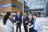 Group of six business professionals having a discussion outside a modern office building with large glass windows and wood paneling.