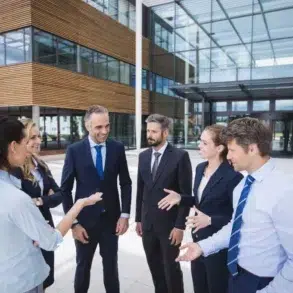 Group of six business professionals having a discussion outside a modern office building with large glass windows and wood paneling.