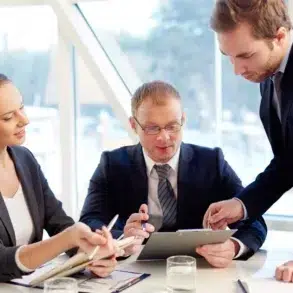 Three business professionals in suits are gathered around a table reviewing documents.