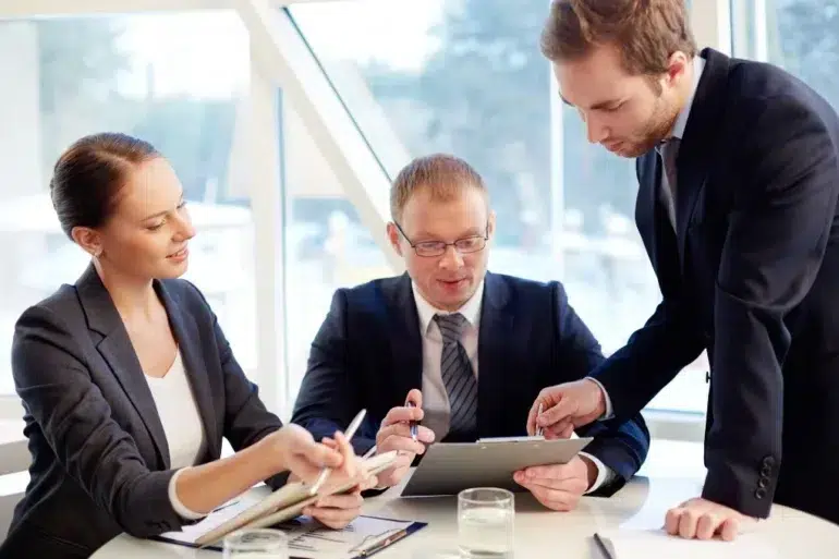 Three business professionals in suits are gathered around a table reviewing documents.