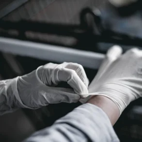 Person wearing white disposable gloves putting on the second glove in a dimly lit environment.