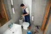A plumber in blue overalls works on a toilet tank in a modern bathroom. Tools are scattered on the floor, conveying a sense of focused repair work.