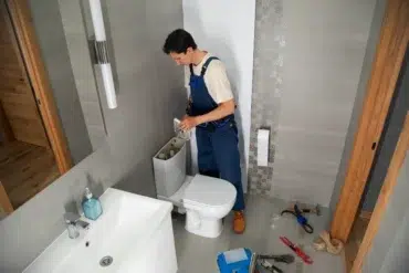 A plumber in blue overalls works on a toilet tank in a modern bathroom. Tools are scattered on the floor, conveying a sense of focused repair work.