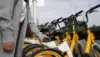 A person stands next to a row of yellow rental bikes, holding a smartphone near a bike's QR code scanner. The scene conveys modern urban commuting.