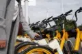 A person stands next to a row of yellow rental bikes, holding a smartphone near a bike's QR code scanner. The scene conveys modern urban commuting.