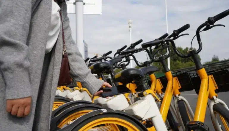 A person stands next to a row of yellow rental bikes, holding a smartphone near a bike's QR code scanner. The scene conveys modern urban commuting.