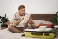 Man sitting cross-legged on bed, checking a list while packing a yellow suitcase with clothes and toiletries.